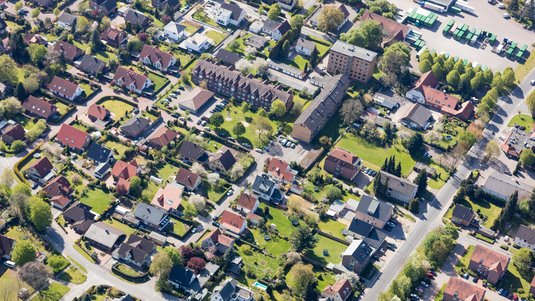 Luftaufnahme eines Wohngebiets mit vielen Einfamilienhäusern, Grünflächen und Straßen in einer sonnigen, dicht bebauten Vorstadt.