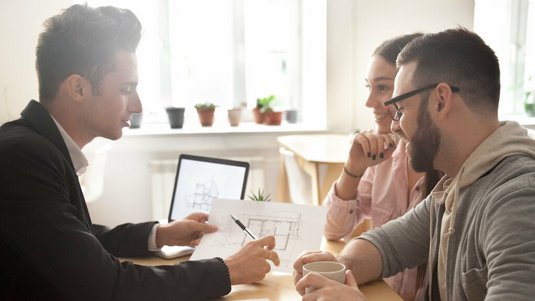 Berater erklärt einem Paar am Tisch einen Hausgrundriss und bespricht Baupläne, während alle entspannt miteinander sprechen.