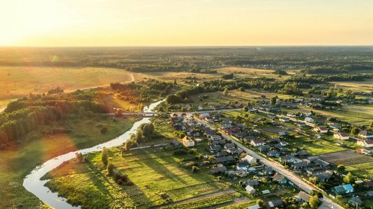 Luftaufnahme einer ländlichen Gegend mit weitläufigen Feldern, einem geschwungenen Fluss, kleinen Siedlungen und sanftem Sonnenlicht über der Landschaft.
