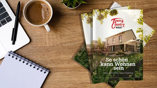 Town-&-Country-Hauskatalog liegt auf einem Schreibtisch neben Laptop, Notizblock und Kaffeetasse; auf dem Titel ist ein modernes Einfamilienhaus abgebildet.