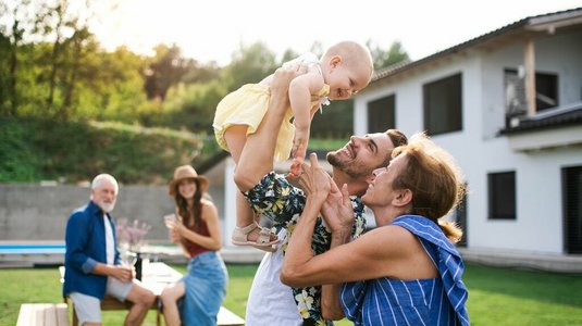 Mehrere Generationen einer Familie verbringen Zeit im Garten eines modernen Hauses; ein Mann hebt ein Baby lachend in die Höhe, während weitere Familienmitglieder im Hintergrund zusammensitzen und sich unterhalten.