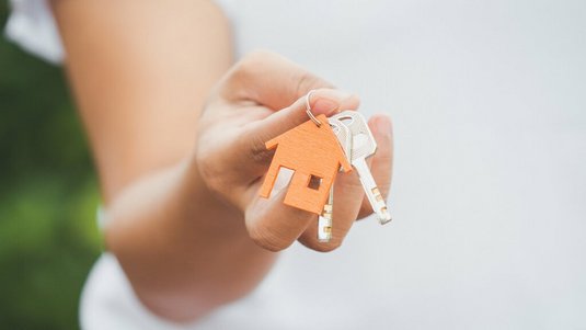 Eine Person hält einen Schlüsselbund mit einem kleinen Hausanhänger in der Hand – Symbol für die Übergabe eines neuen Eigenheims.