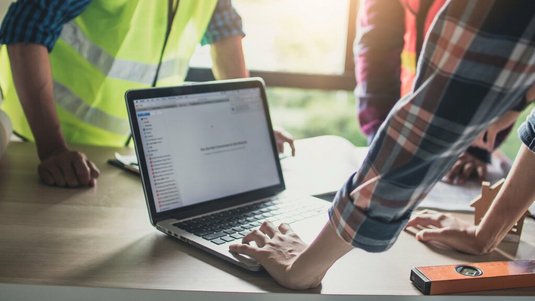 Mehrere Personen in Arbeitskleidung stehen an einem Tisch und prüfen gemeinsam Unterlagen und Daten auf einem Laptop, daneben liegen Baupläne und ein Messwerkzeug.