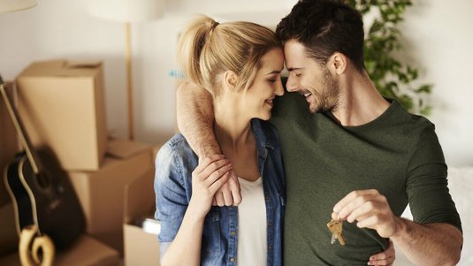 Glückliches Paar steht in einem neuen Zuhause zwischen Umzugskartons; eine Person hält einen Schlüssel in der Hand.