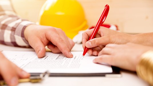 Zwei Personen prüfen und unterschreiben ein Bau- oder Vertragsdokument auf einem Tisch; im Hintergrund liegen ein Stift und ein gelber Bauhelm.