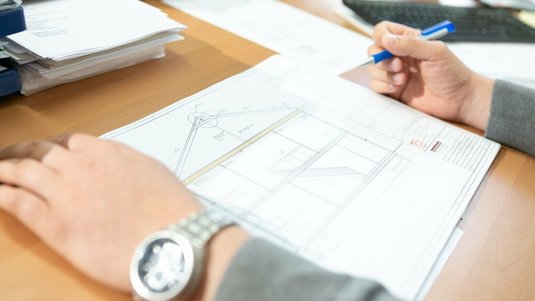 Person betrachtet und markiert Bauzeichnungen und Grundrisspläne auf einem Schreibtisch, umgeben von Unterlagen und Schreibmaterial.