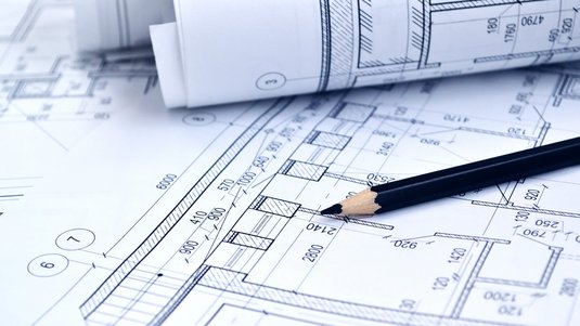 Detailaufnahme eines Bauplans mit eingezeichneten Grundrissen, Maßangaben und einem Bleistift, der auf einen Raum zeigt.