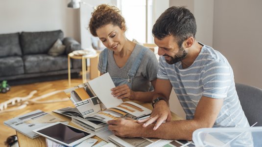 Zwei Personen sitzen in einem Wohnzimmer am Tisch und blättern gemeinsam in Wohn-/Designkatalogen und Musterunterlagen.