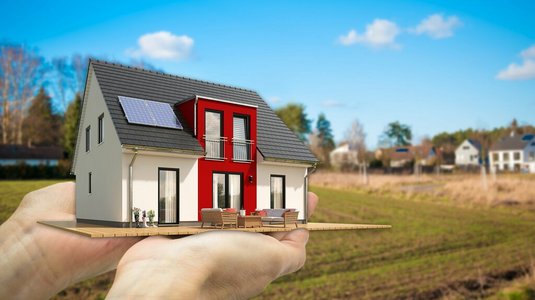 Hände halten ein Modell eines modernen Einfamilienhauses mit Solarpanel vor einer freien Grundstücksfläche in ländlicher Umgebung.
