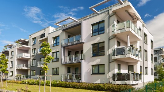 Modernes, helles Mehrfamilienhaus mit mehreren Glasbalkonen, großen Fenstern und begrüntem Außenbereich unter blauem Himmel.