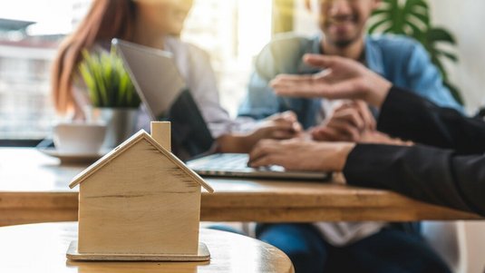 Kleines Holzhausmodell steht auf einem Tisch im Vordergrund, während im Hintergrund mehrere Personen in einem Beratungsgespräch zusammensitzen.