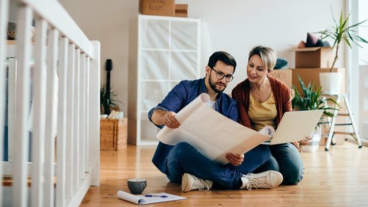 Paar sitzt auf dem Boden eines neuen Zuhauses und betrachtet gemeinsam Baupläne und Unterlagen, während Umzugskartons und eine Leiter im Hintergrund stehen.