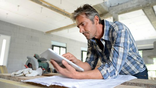 Mann auf einer Baustelle prüft Bauunterlagen auf einem Tablet, während Pläne und Werkzeuge auf dem Tisch liegen.