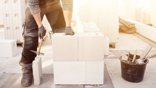 Handwerker mit Arbeitshandschuhen setzt weiße Porenbetonsteine zu einer Mauer zusammen; daneben liegen Werkzeug und ein Eimer auf der Baustelle im Gegenlicht.