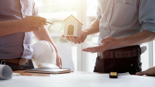 Zwei Personen stehen an einem Tisch mit Bauplänen; eine hält ein Hausmodell, die andere zeigt mit einem Stift darauf, während ein Bauhelm und Unterlagen bereitliegen.