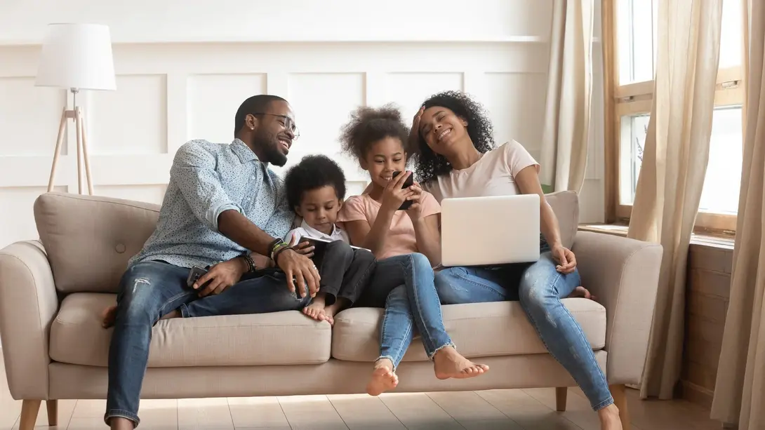 Familie sitzt entspannt auf dem Sofa und nutzt gemeinsam Smartphone und Laptop im Wohnzimmer.