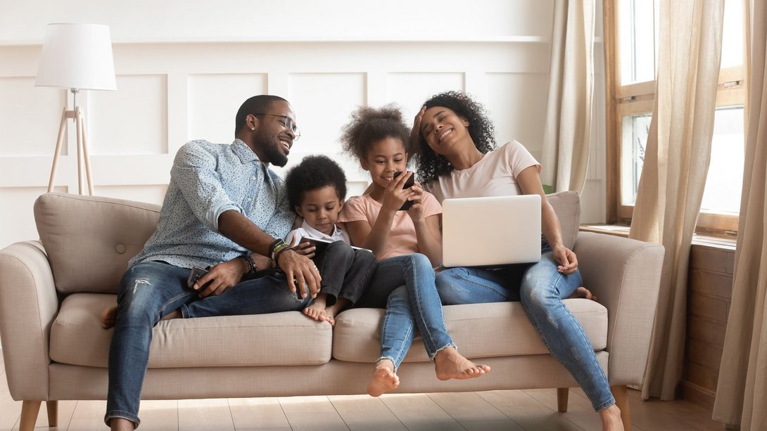 Familie sitzt entspannt auf dem Sofa und nutzt gemeinsam Smartphone und Laptop im Wohnzimmer.
