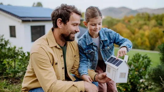 Vater und Kind halten ein Hausmodell mit Solarpanel auf dem Dach im Garten vor einem modernen Haus.