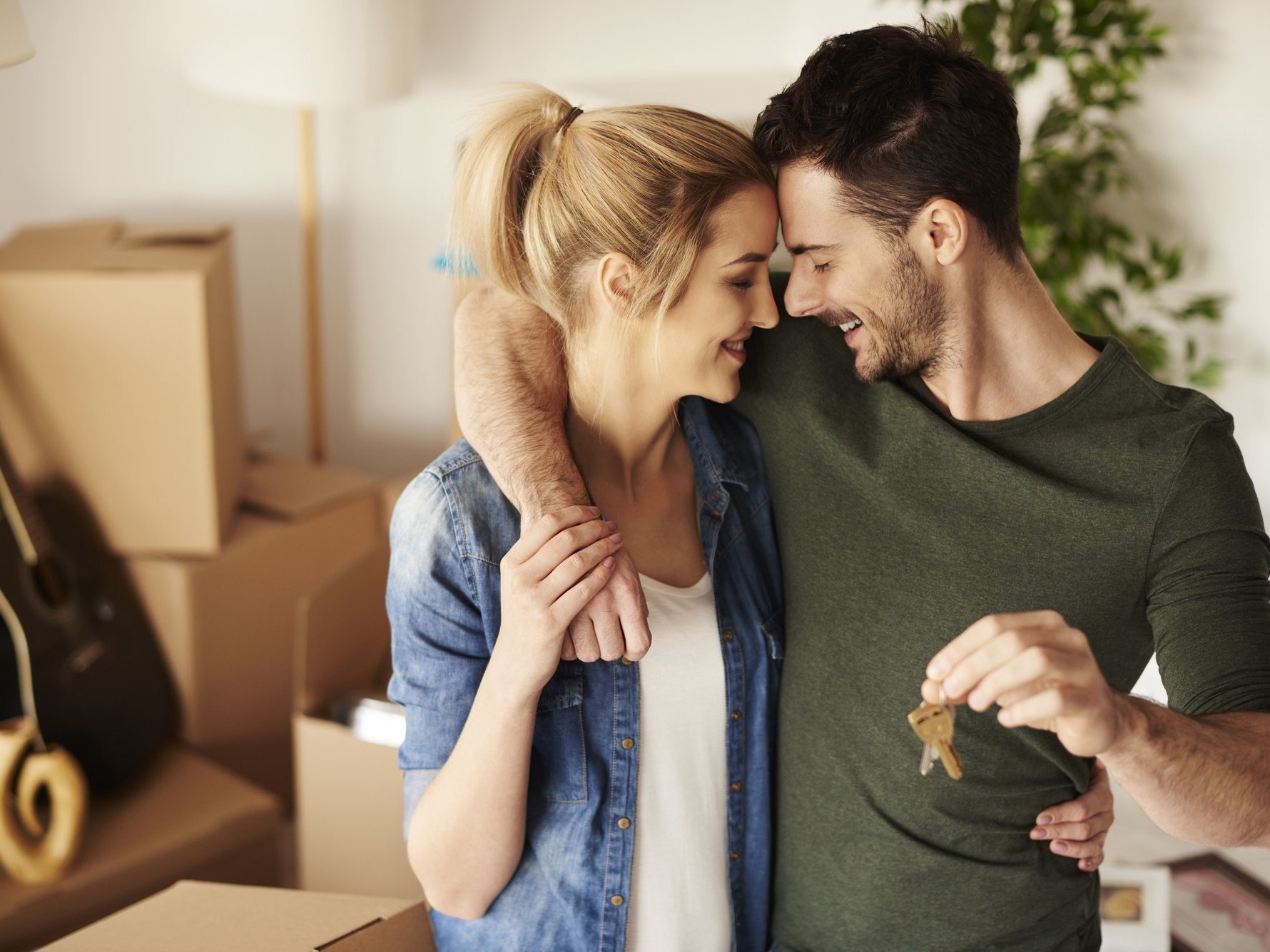 Junges Paar steht lächelnd in einer Wohnung mit Umzugskartons, der Mann hält einen Schlüssel in der Hand, beide umarmen sich.