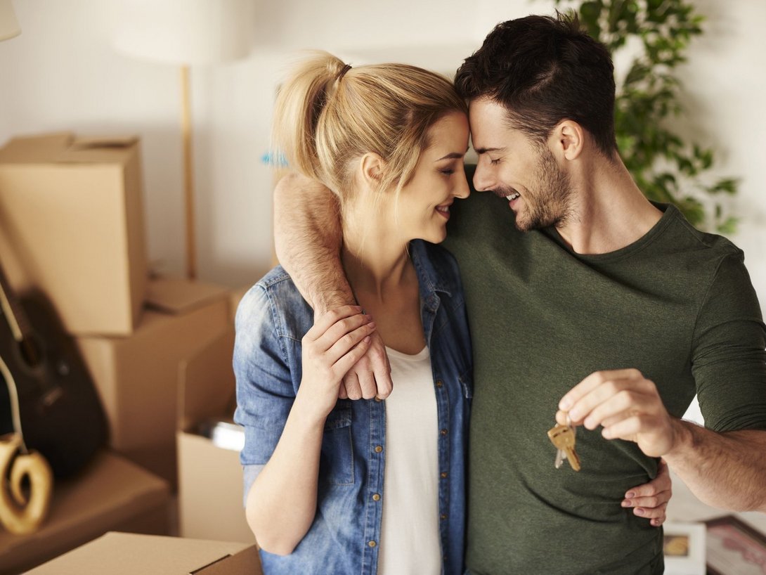 Junges Paar steht lächelnd in einer Wohnung mit Umzugskartons, der Mann hält einen Schlüssel in der Hand, beide umarmen sich.