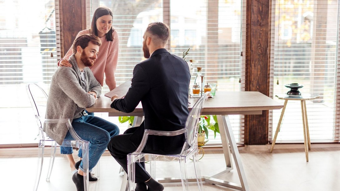Junges Paar sitzt mit einem Finanzberater am Tisch und bespricht Unterlagen zur Baufinanzierung.
