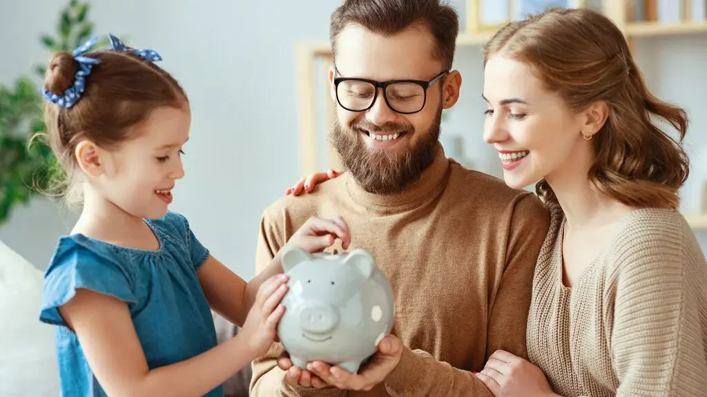Haus-Familie-Wohnen-Serie-19-15 Familie mit Eltern und Kind hält gemeinsam ein Sparschwein und legt Münzen hinein – Symbol für gemeinsames Sparen und finanzielle Planung.