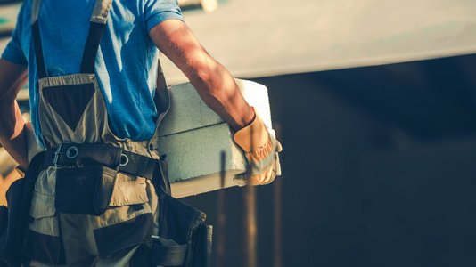Bauarbeiter in Arbeitskleidung trägt einen Betonstein auf einer Baustelle; der Hintergrund zeigt im Bau befindliche Strukturen.