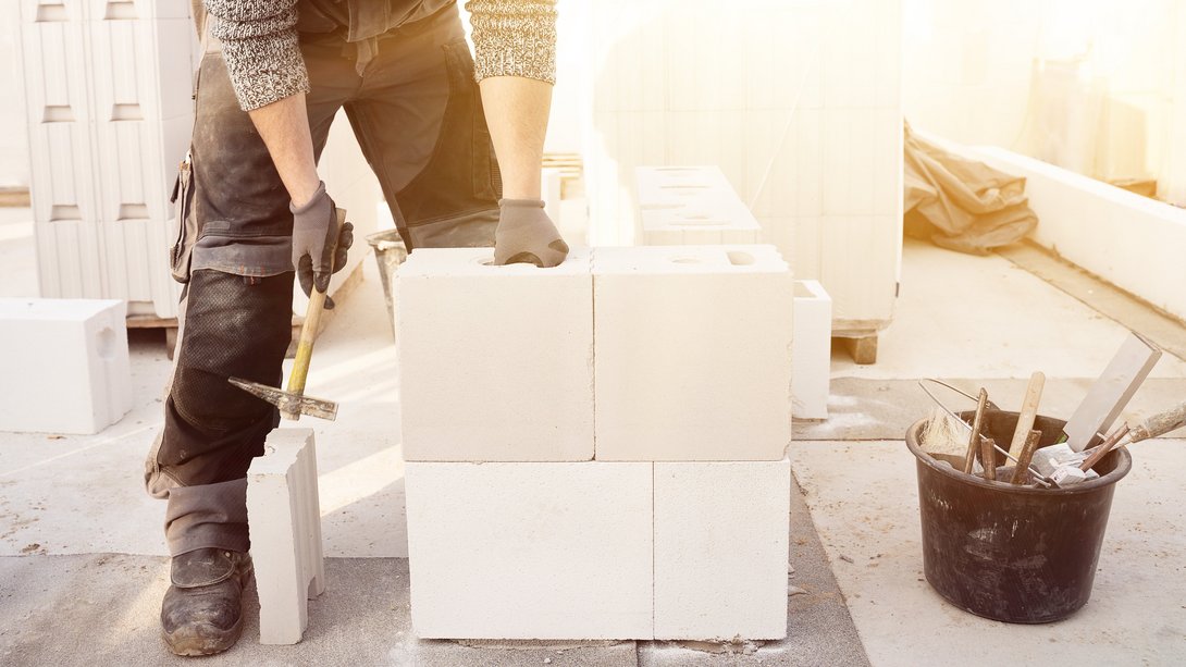 Klimagerecht-Bauen-16 Handwerker mit Arbeitshandschuhen setzt weiße Porenbetonsteine zu einer Mauer zusammen; daneben liegen Werkzeug und ein Eimer auf der Baustelle im Gegenlicht.