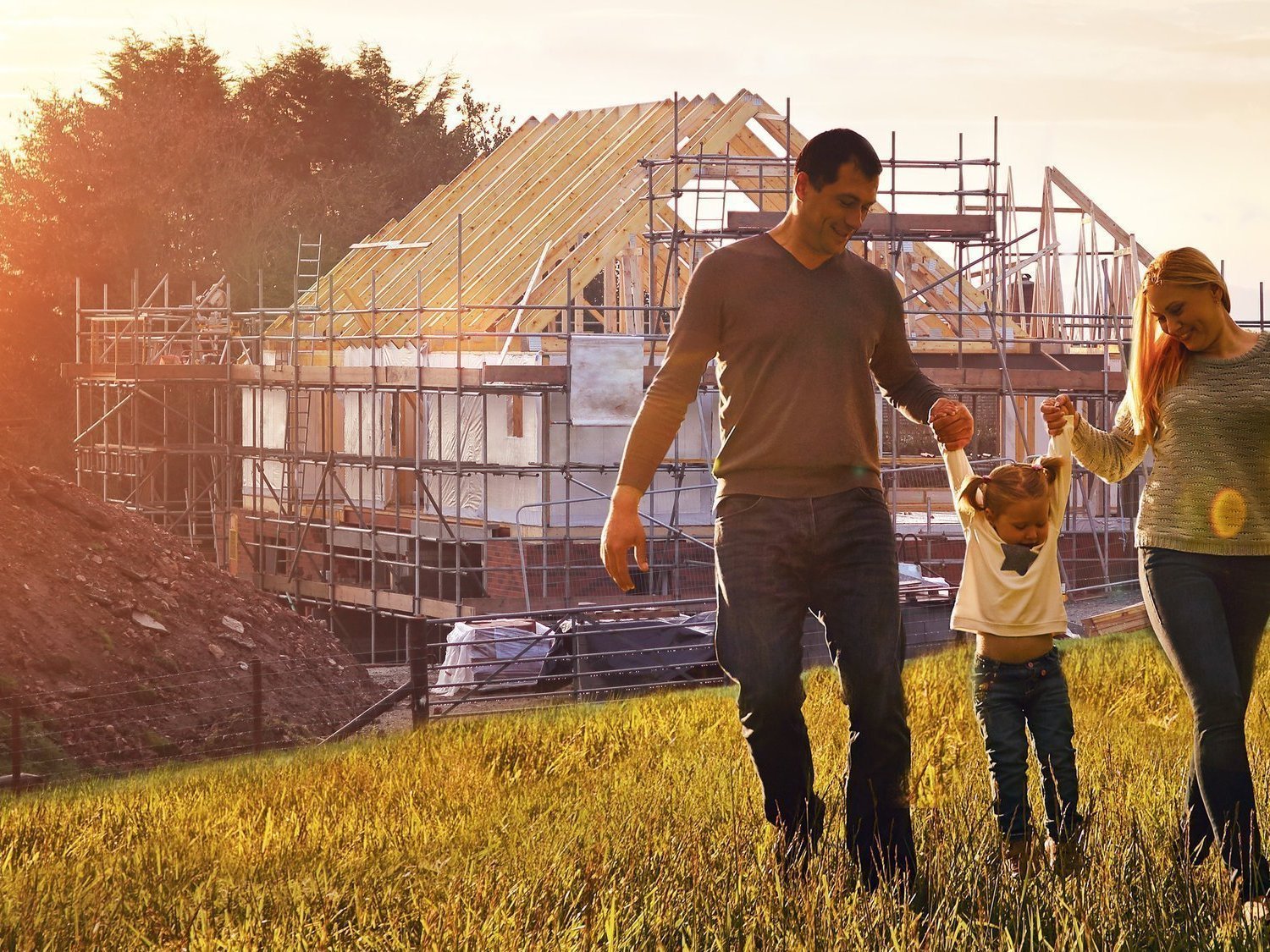 Familie mit kleinem Kind läuft über eine Wiese, im Hintergrund steht ein Einfamilienhaus im Rohbau mit Dachkonstruktion und Baugerüst.