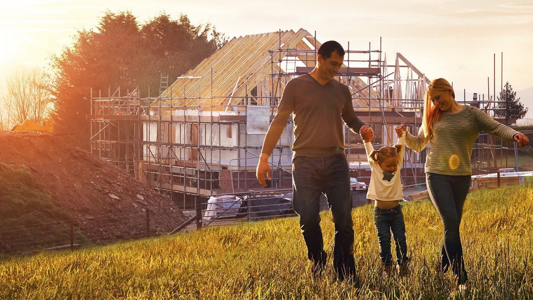Familie mit Kind spaziert über ein Grundstück vor einem im Bau befindlichen Haus mit Gerüst bei Sonnenuntergang.