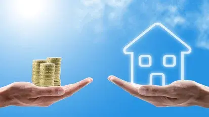 Zwei Hände halten links einen Stapel Goldmünzen und rechts ein leuchtendes Haus-Symbol vor blauem Himmel.