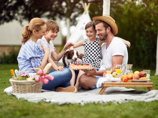 Familie mit zwei Kindern sitzt auf einer Picknickdecke im Garten, isst gemeinsam und spielt mit einem Hund bei sonnigem Wetter.