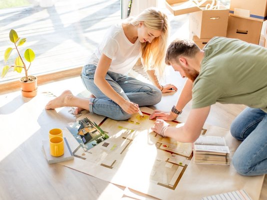 Frau und Mann sitzen auf dem Boden einer hellen Wohnung und betrachten gemeinsam ausgebreitete Baupläne und Grundrisse.