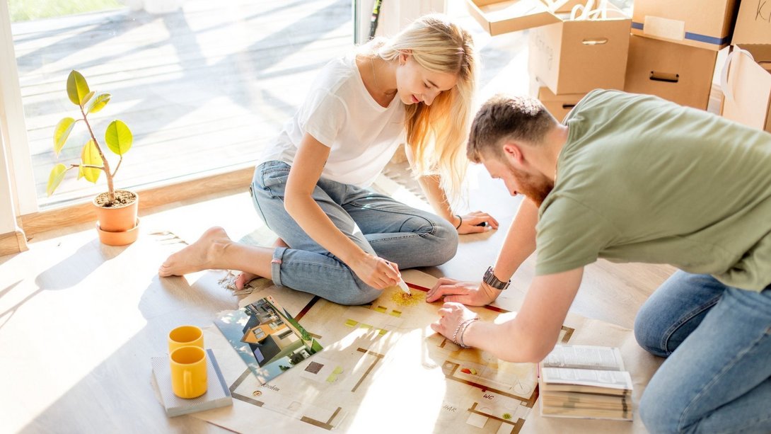 Junges Paar sitzt auf dem Boden und plant gemeinsam den Grundriss ihres zukünftigen Hauses anhand von Bauplänen.