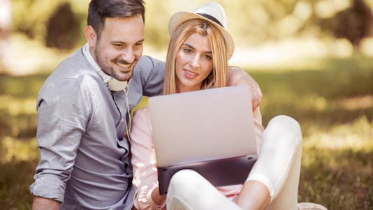 Junges Paar sitzt entspannt im Park und schaut gemeinsam auf einen Laptop – Symbol für digitale Finanz- und Budgetplanung.