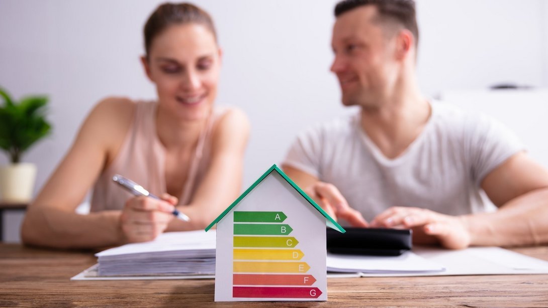 Paar sitzt am Tisch und plant die Finanzierung, während im Vordergrund ein Hausmodell mit Energieeffizienzskala (A–G) steht.