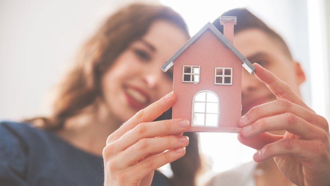 Junges Paar hält gemeinsam ein kleines Modellhaus in den Händen und lächelt.