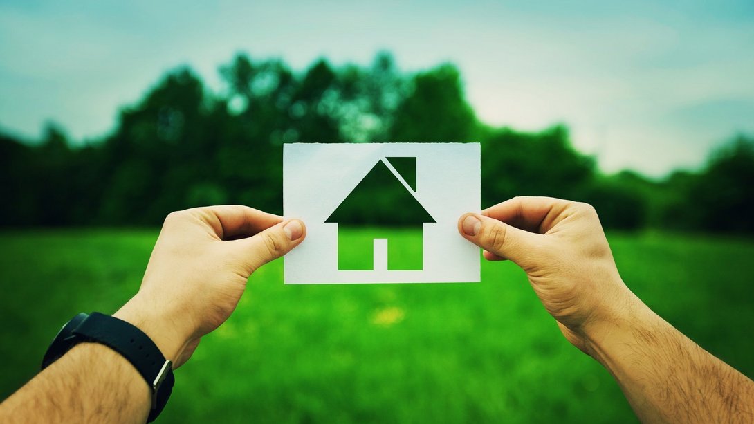 Hände halten eine Karte mit ausgeschnittenem Haus-Symbol vor einer grünen Wiese – Sinnbild für Grundstück und Hausbauplanung.