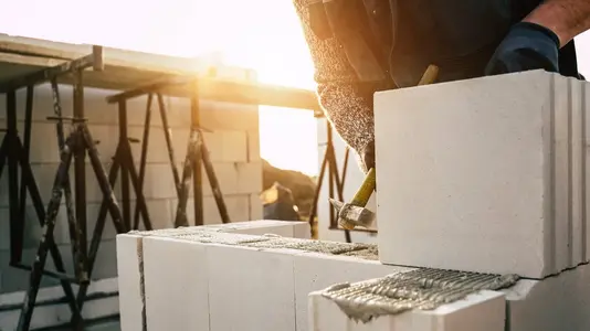 Handwerker setzt große Porenbetonsteine auf frischen Mörtel und richtet sie beim Bau einer Massivhauswand im Sonnenlicht aus.