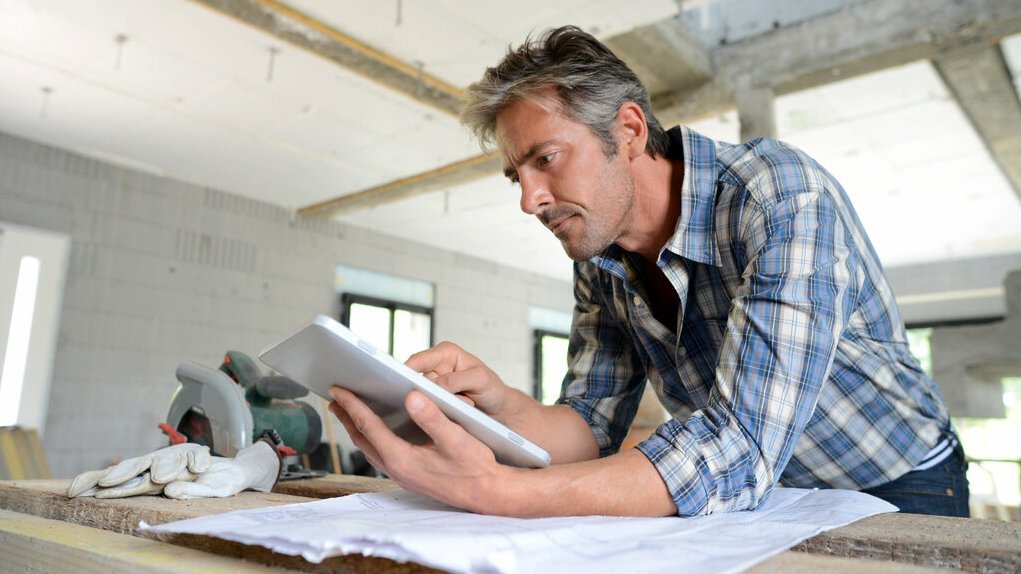 hausbau-haus-bauen-eigenleistung-handwerker-rohbau-01 Mann auf einer Baustelle prüft Bauunterlagen auf einem Tablet, während Pläne und Werkzeuge auf dem Tisch liegen.