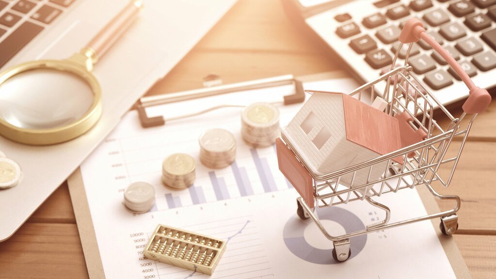 finanzierung-vertrag-termin-gespraech-unterlagen-13 Miniaturhaus im Einkaufswagen auf Finanzunterlagen mit Diagrammen, Münzen, Taschenrechner und Lupe auf einem Schreibtisch.