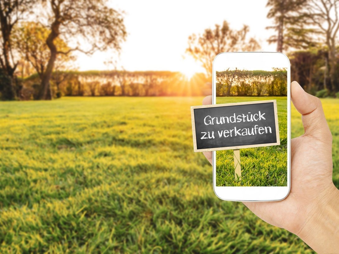 Hand hält ein Smartphone vor einer grünen Wiese mit einem kleinen Schild mit der Aufschrift „Grundstück zu verkaufen“.
