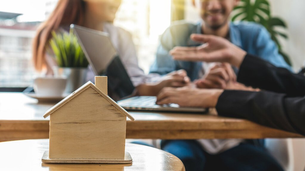 verkauf-verkaufsgespraech-beratung-bauberater-21 Kleines Holzhausmodell steht auf einem Tisch im Vordergrund, während im Hintergrund mehrere Personen in einem Beratungsgespräch zusammensitzen.