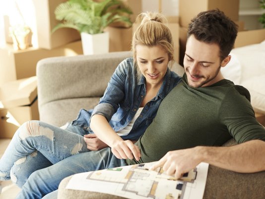 Paar sitzt auf einem Sofa und betrachtet gemeinsam einen Grundrissplan eines Hauses auf einem Tablet.