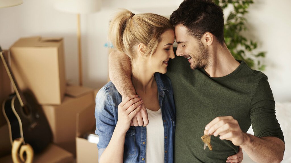 Haus-Paar-Umzug-Serie-13-20 Glückliches Paar steht in einem neuen Zuhause zwischen Umzugskartons; eine Person hält einen Schlüssel in der Hand.