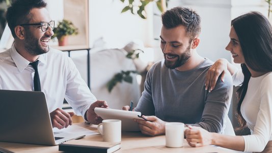 verkauf-verkaufsgespraech-beratung-bauberater-03 Berater sitzt mit einem Paar an einem Tisch und erklärt Unterlagen auf einem Tablet; Laptop, Dokumente und Kaffeetassen stehen auf dem Tisch.