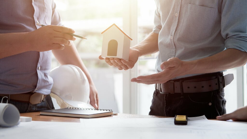 finanzierung-vertrag-termin-gespraech-unterlagen-03 Zwei Personen stehen an einem Tisch mit Bauplänen; eine hält ein Hausmodell, die andere zeigt mit einem Stift darauf, während ein Bauhelm und Unterlagen bereitliegen.