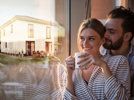 Paar steht am Fenster mit einer Tasse Kaffee und blickt nach draußen; im Spiegelbild ist ein modernes Einfamilienhaus zu sehen.