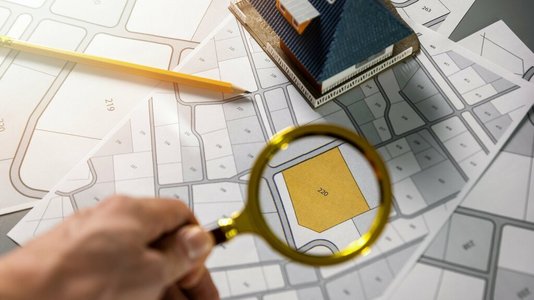 grundstueck-bebauungsplan-grundstueckssuche-05 Hand hält eine Lupe über einen Bebauungsplan und hebt ein markiertes Grundstück hervor; daneben liegen ein Bleistift und ein Hausmodell.