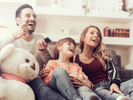 Familie mit Vater, Mutter und Kind sitzt lachend auf einem Sofa im Wohnzimmer und schaut gemeinsam fern.